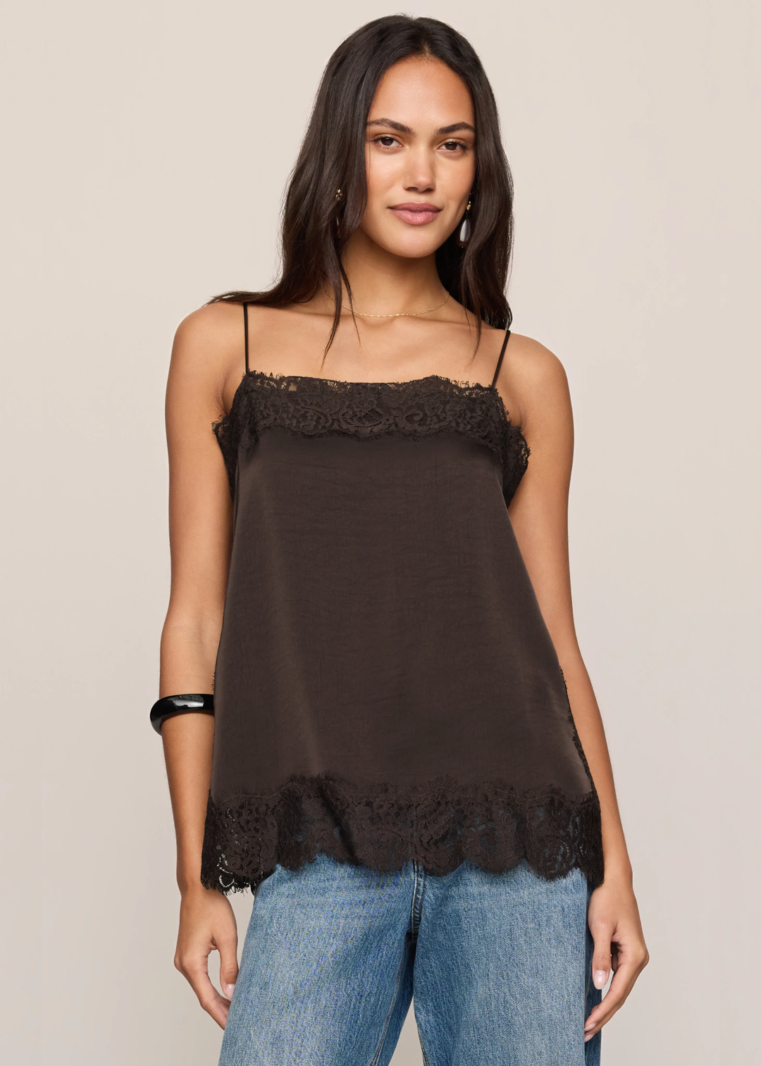A woman modeling a dark brown camisole top with delicate lace trim along the neckline and hem, paired with blue jeans, against a solid light beige background.