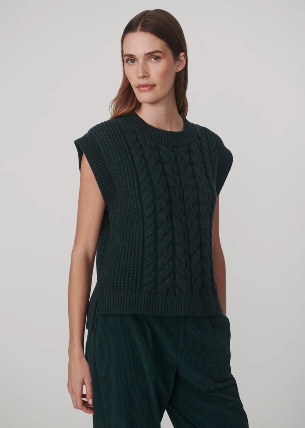 A woman modeling a dark forest green sleeveless cable-knit sweater vest with a crew neck, paired with matching dark green trousers, against a light gray studio background.