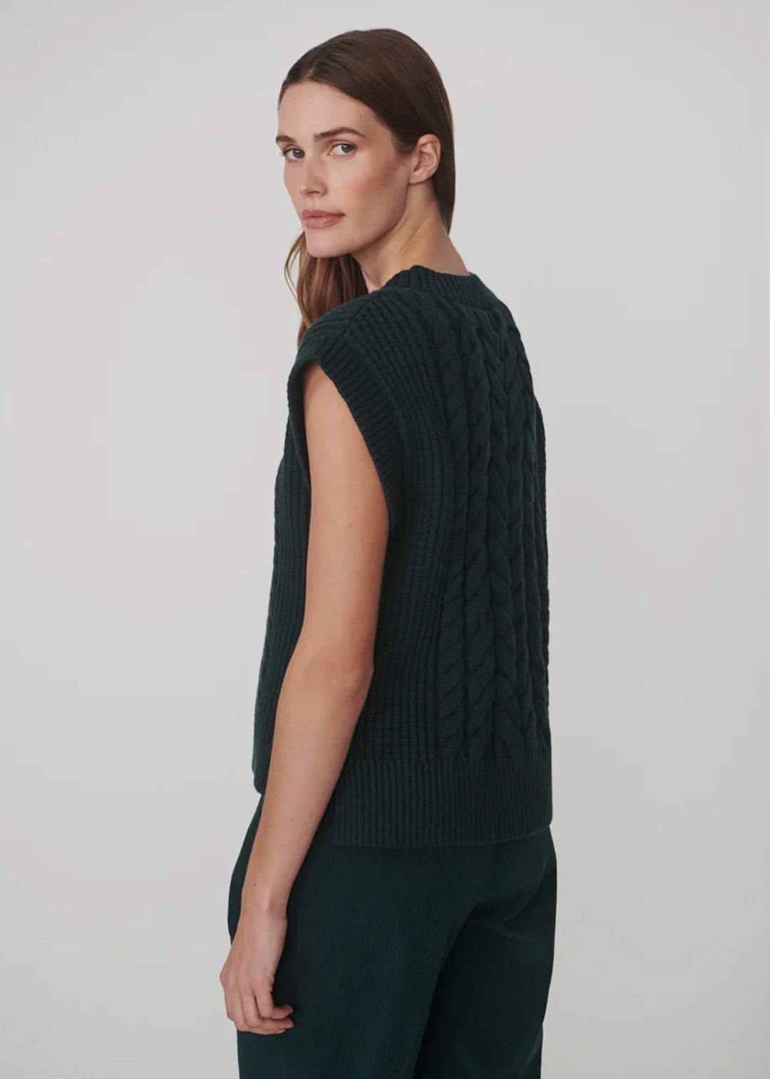 A woman shown from the back wearing a dark green sleeveless cable-knit sweater vest and matching dark green trousers, photographed against a plain light gray studio background.