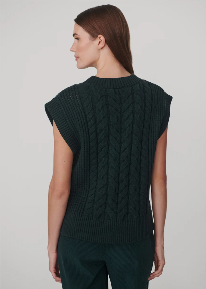 A woman shown from the back wearing a dark green cable-knit sleeveless sweater vest and matching dark green trousers, against a plain light gray studio background.