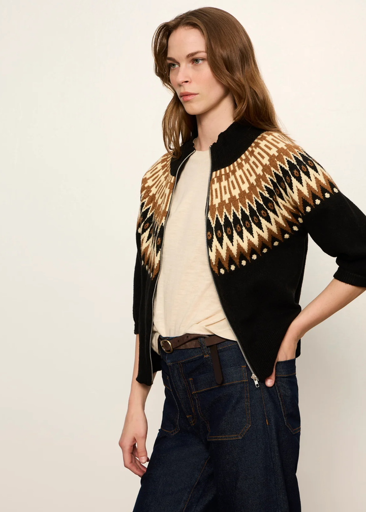 A woman modeling a black knit cardigan with a geometric Fair Isle pattern in beige, brown, and black across the shoulders and upper chest, styled with a cream t-shirt and dark blue jeans against a plain off-white background.
