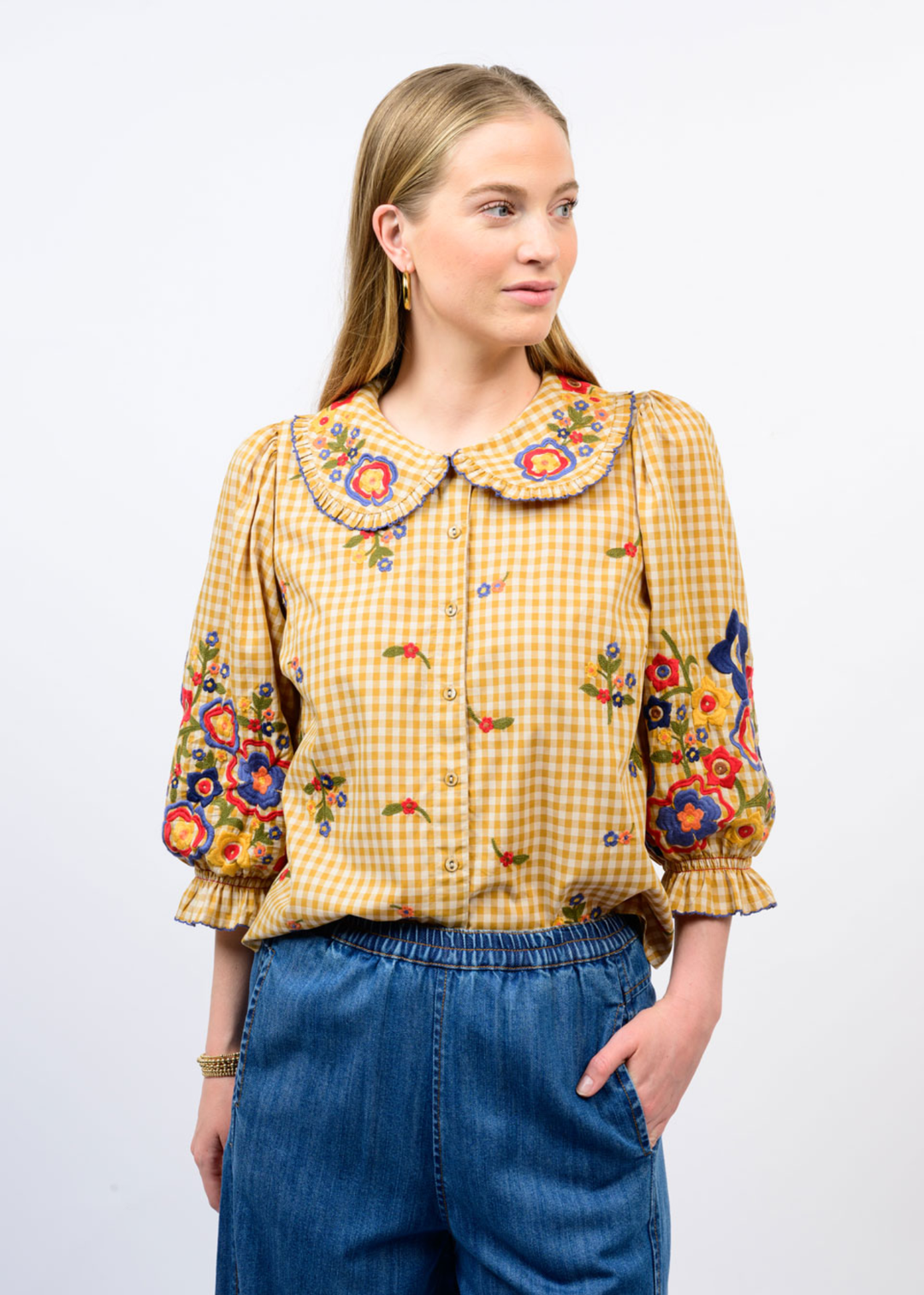 A woman modeling a yellow and white gingham blouse with colorful floral embroidery on the collar, cuffs, and body, paired with blue jeans against a plain white background.