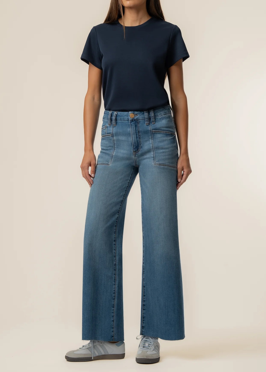 A model wearing a navy blue short-sleeve t-shirt, high-waisted wide-leg blue denim jeans, and gray sneakers, standing against a plain beige studio background.