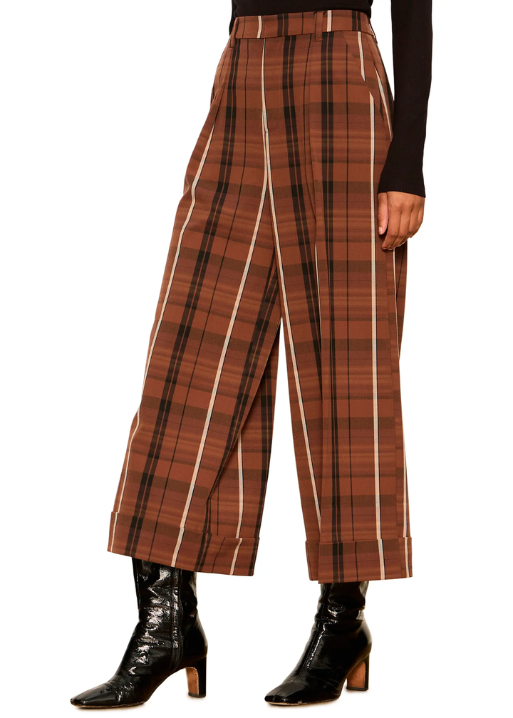 A model is wearing high-waisted, wide-leg cropped trousers in a brown and black plaid pattern with vertical white stripe accents, styled with black pointed-toe ankle boots on a white background.