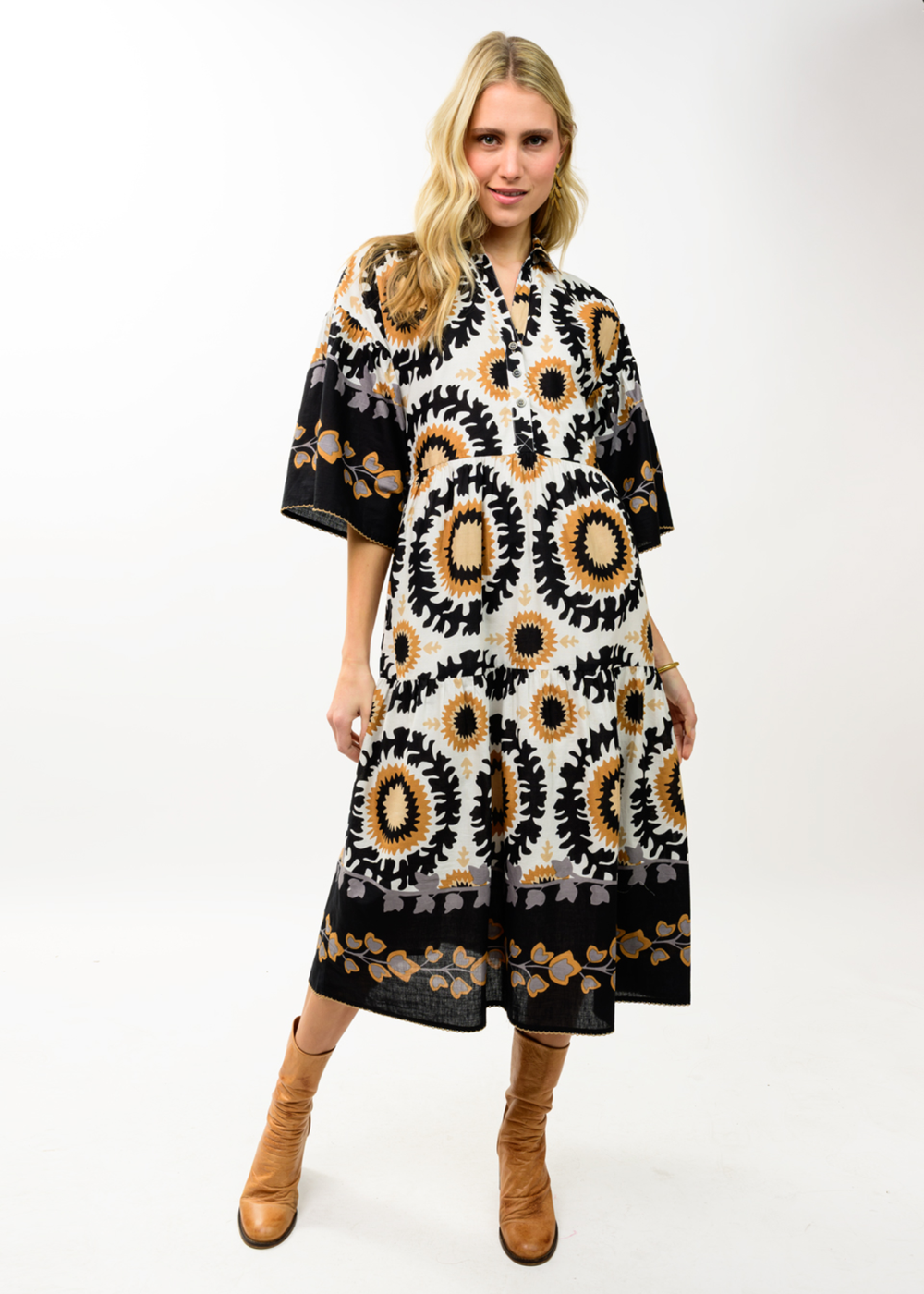 A woman modeling a loose, midi-length dress with a black, white, and tan circular sunflower pattern, paired with tan boots, against a plain white studio background.