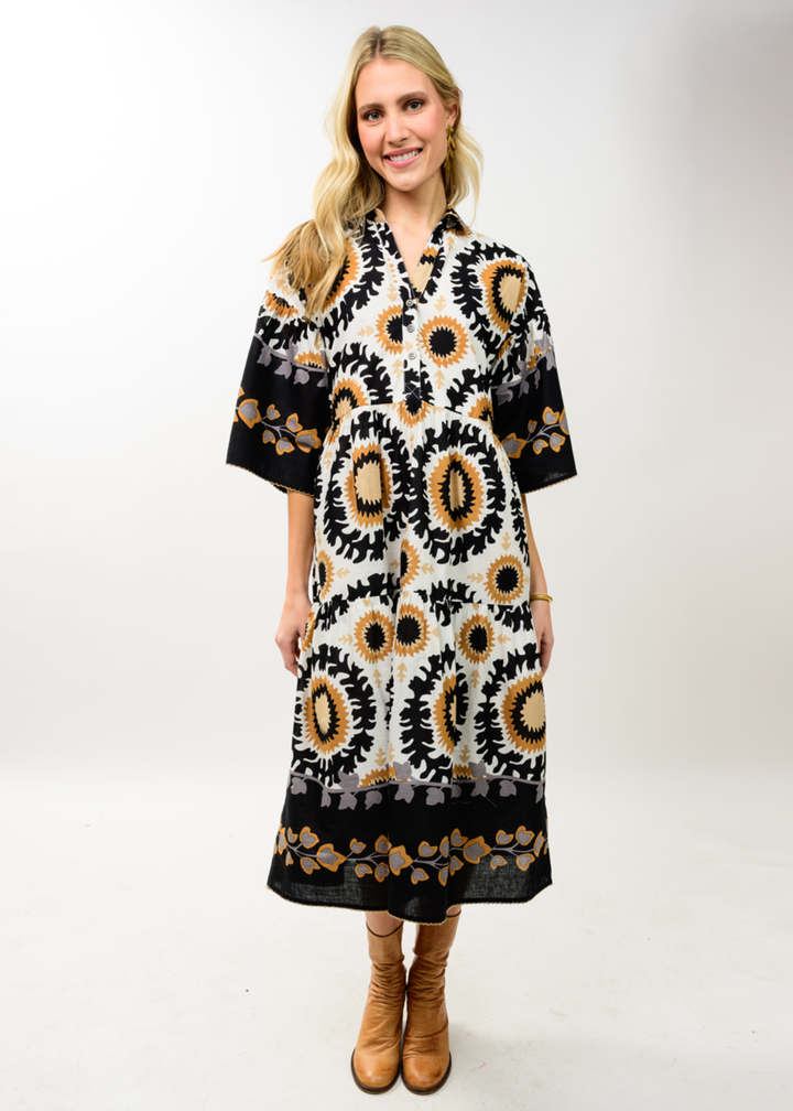 A woman modeling a black, white, and tan kaftan dress with circular sunburst patterns, paired with tan ankle boots, against a plain white studio background.