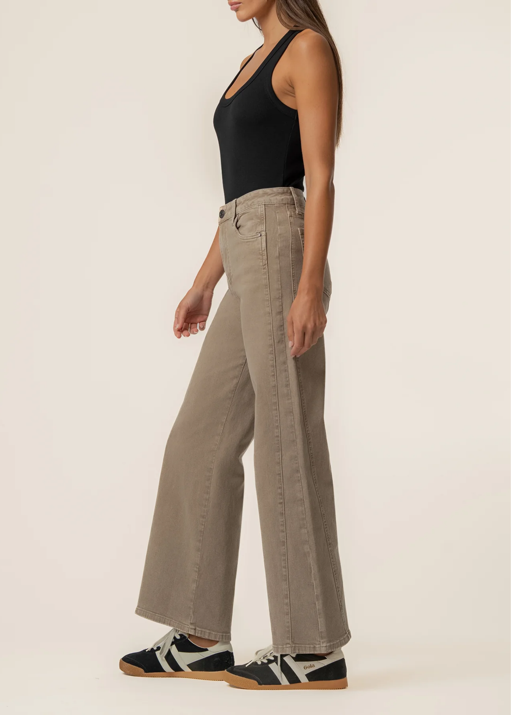 A woman modeling a pair of wide-leg beige jeans with a black tank top and black-and-white sneakers, shown from the side against a plain off-white studio background.