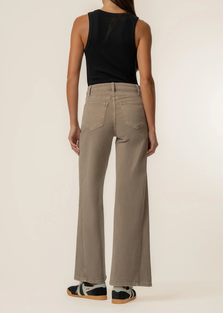 A model shown from the back wearing high-waisted, wide-leg taupe pants, a black sleeveless top, and black-and-white sneakers against a plain off-white studio background.