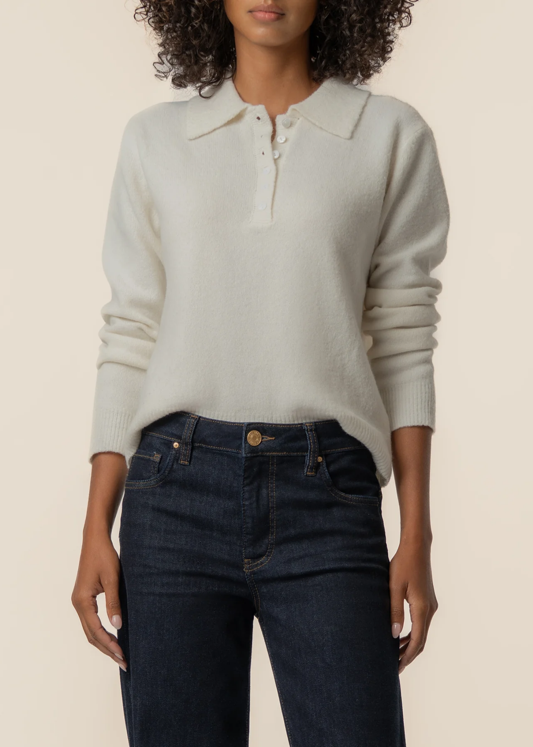 A woman wearing a cream-colored long-sleeve knit polo sweater and dark blue jeans, shown from the neck down against a solid light beige background.
