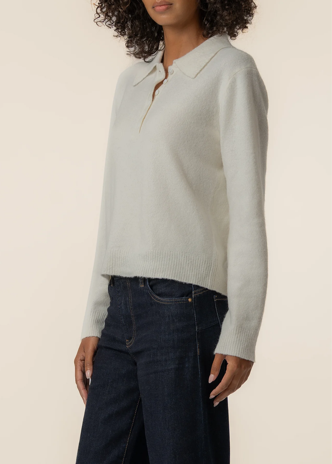 A woman wearing a light cream knit polo sweater and dark blue jeans, shown from the side against a plain beige background.