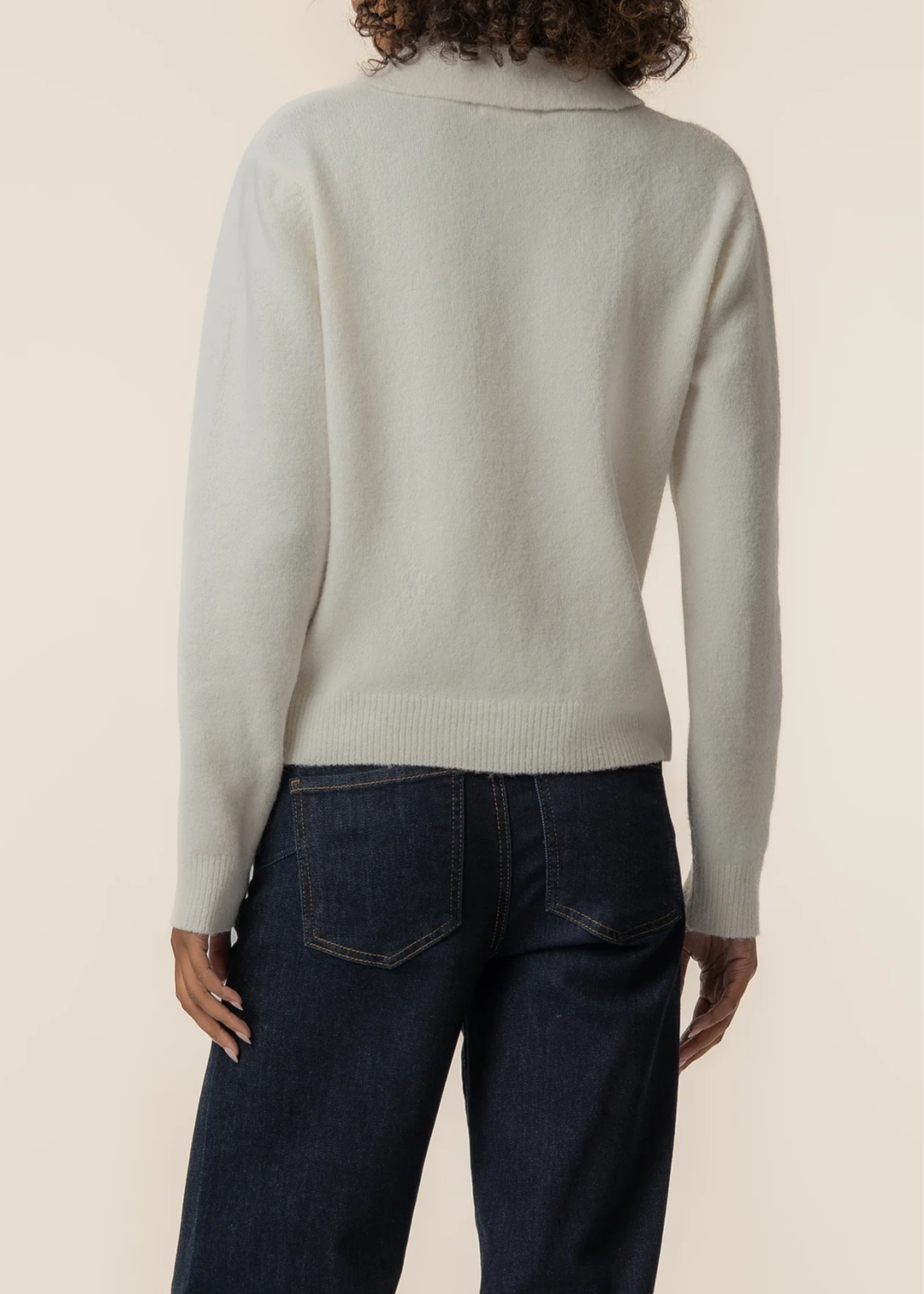 A person shown from the back wearing a light cream knit sweater and dark blue jeans against a plain beige studio background.