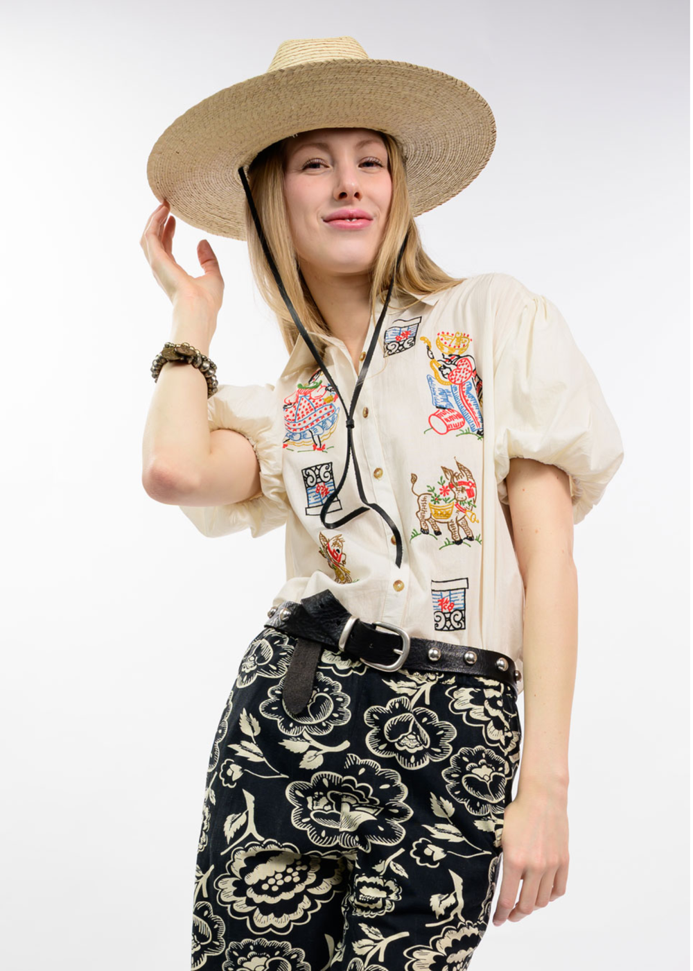 A woman wearing a straw cowboy hat, a cream short-sleeve button-up shirt with colorful embroidered patches, and black floral pants with a black belt, posed against a light gray studio background.