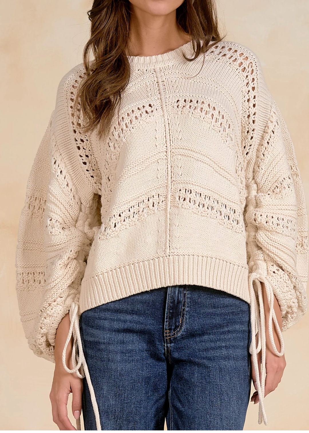 A woman wearing a cream-colored, chunky open-knit sweater with balloon sleeves and side tie details, paired with blue jeans, against a warm beige background.