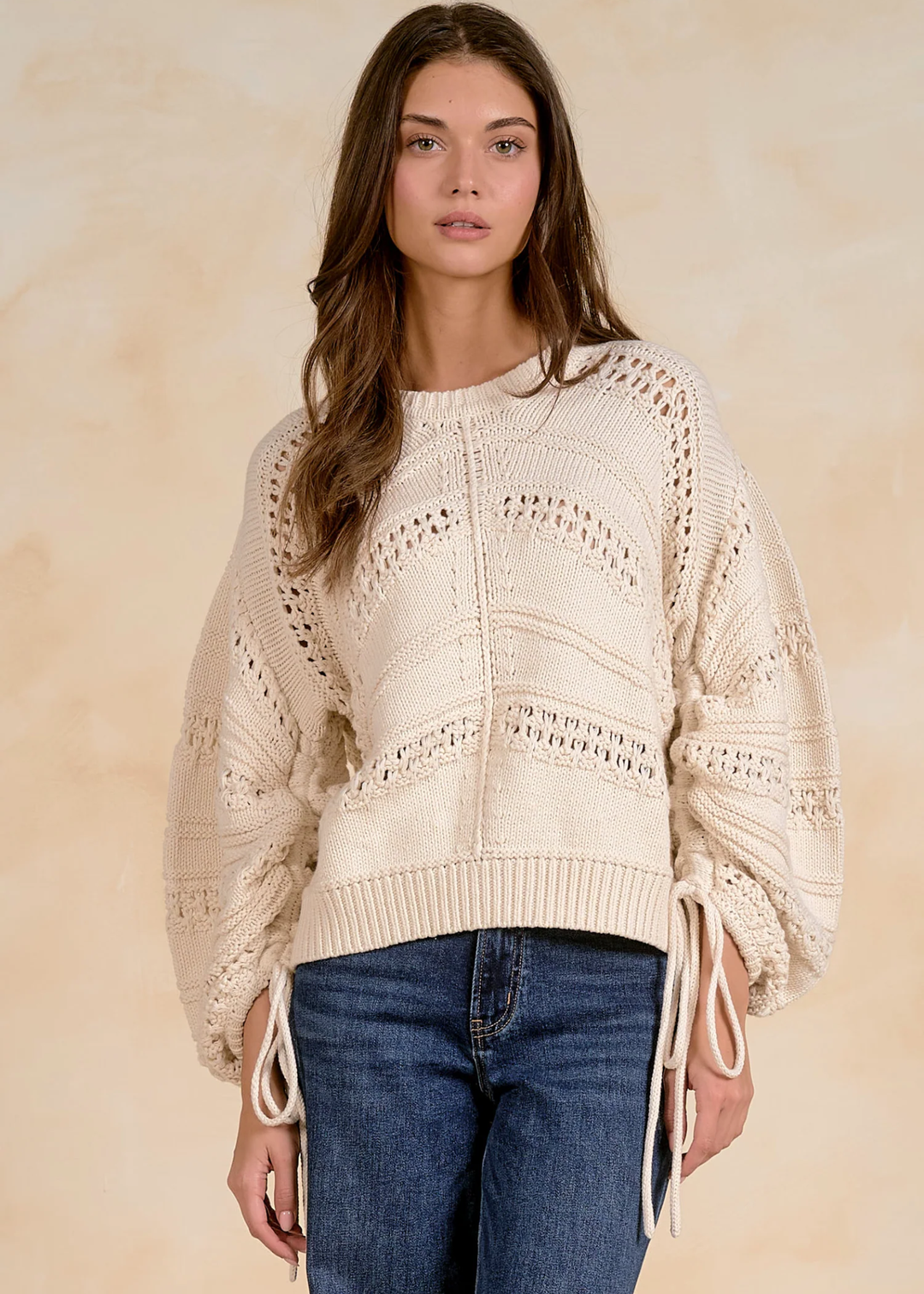 A woman wearing a cream-colored, textured knit sweater with balloon sleeves and tie details at the cuffs, paired with blue jeans, set against a warm beige background.