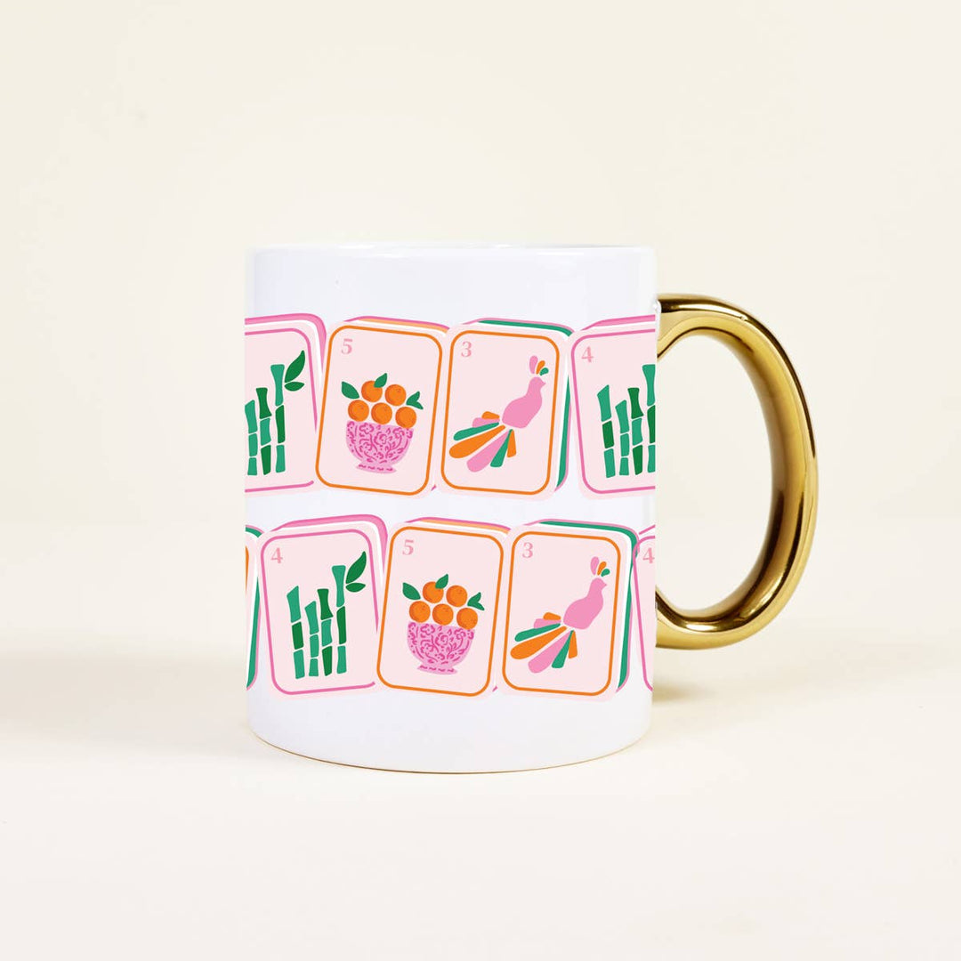 A white ceramic mug with a gold handle, decorated with a pattern of pink and orange playing cards featuring bamboo, peacocks, and fruit. The mug is photographed on a clean, solid, warm off-white background.