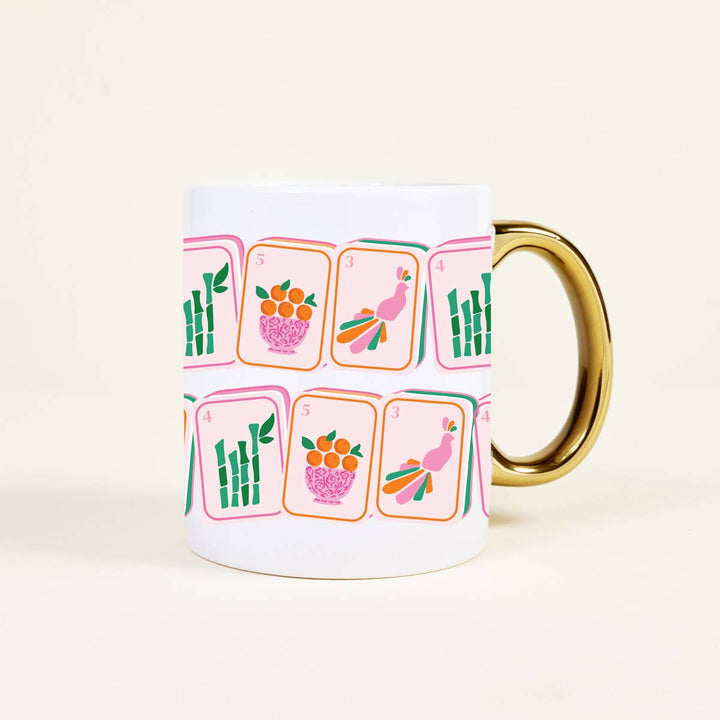 A white ceramic mug with a gold handle, decorated with a pattern of pink and orange playing cards featuring bamboo, peacocks, and fruit. The mug is photographed on a clean, solid, warm off-white background.