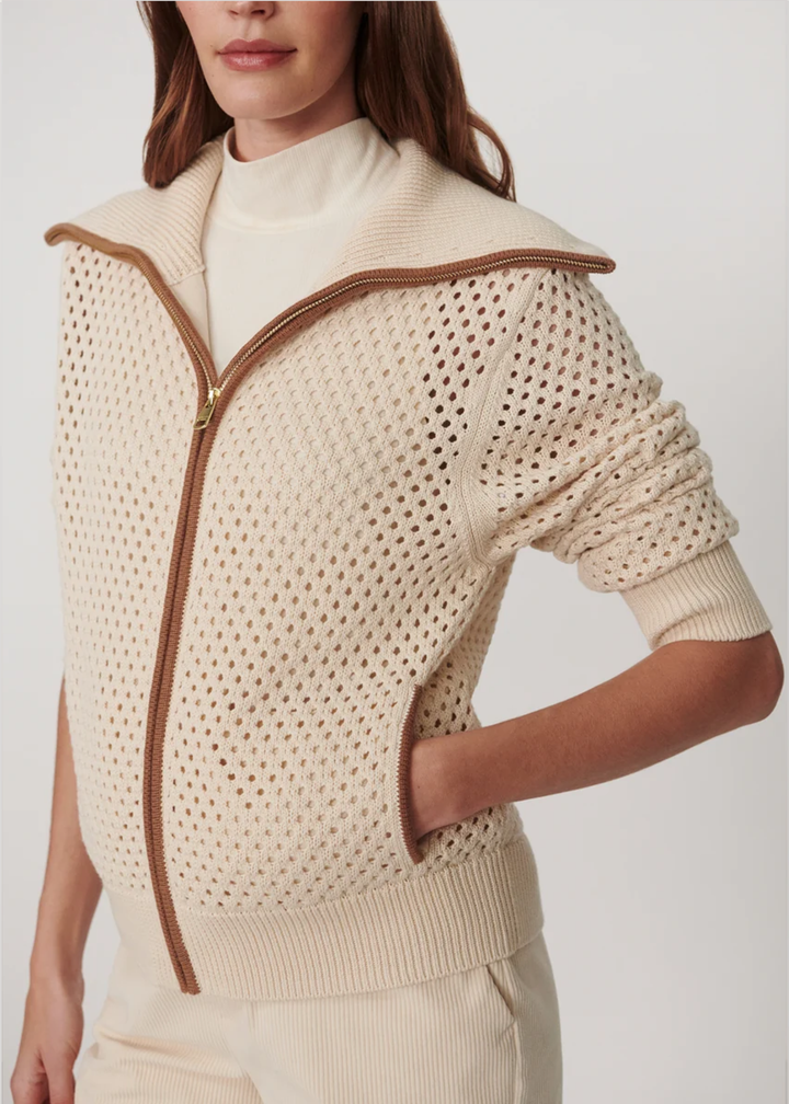 A close-up of a model wearing a cream-colored open-knit zip-up sweater with brown trim and a high collar, paired with a cream mock neck top and cream pants against a light gray studio background.