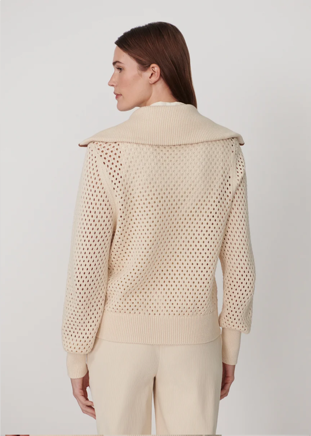 A woman shown from the back wearing a cream-colored knit sweater with a wide collar and openwork knit pattern, paired with matching cream pants, against a plain light gray studio background.
