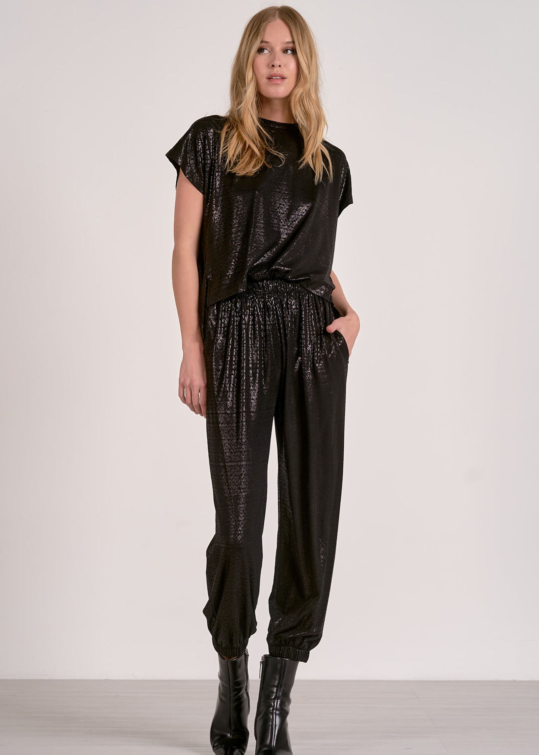 Woman wearing a shiny black short sleeve jumpsuit against a grey wall