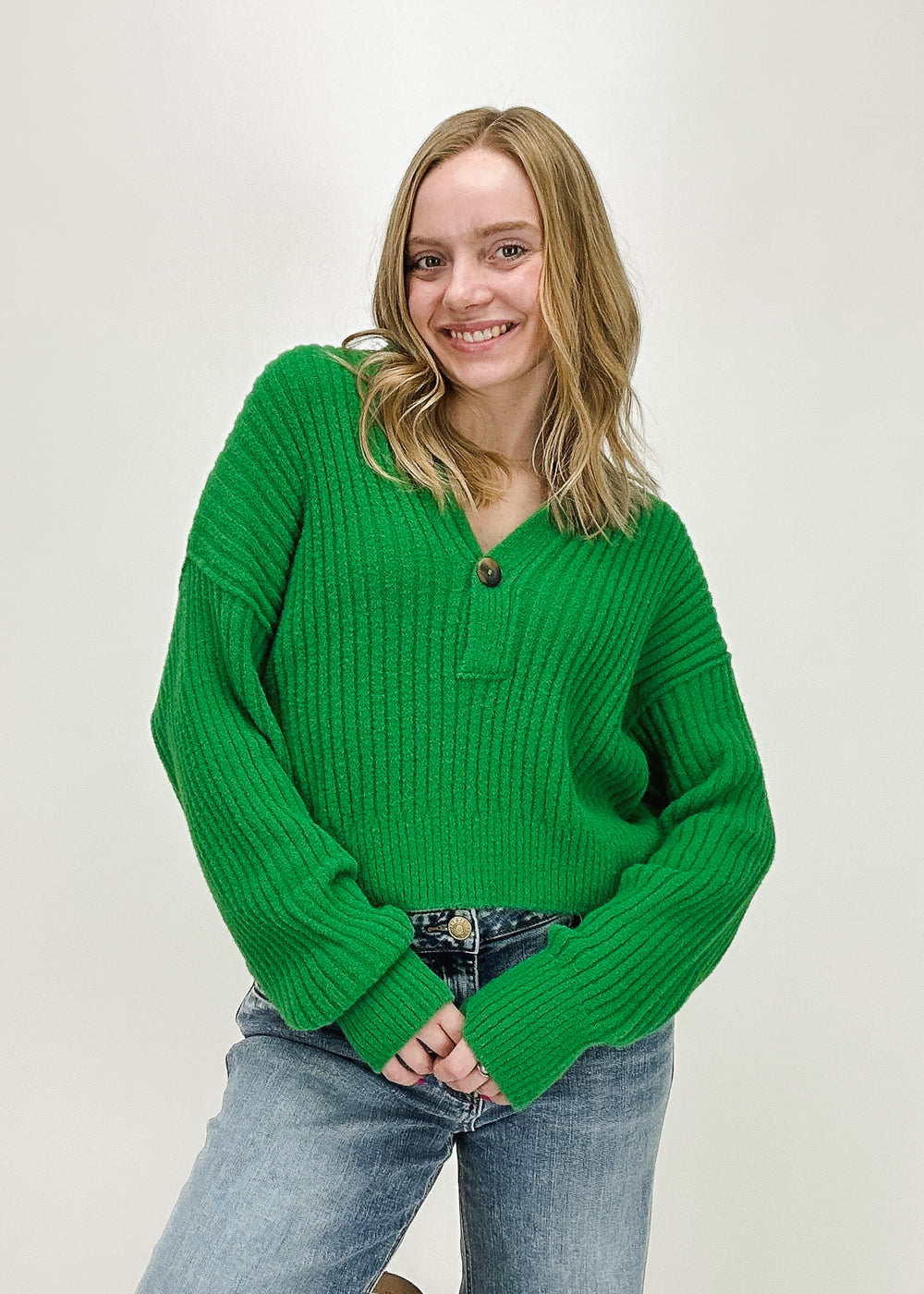 A woman wearing the Free People Cocoa Henley in Rolling Hills, a green ribbed V-neck sweater with a button detail, styled with light-wash jeans for a cozy yet stylish look.