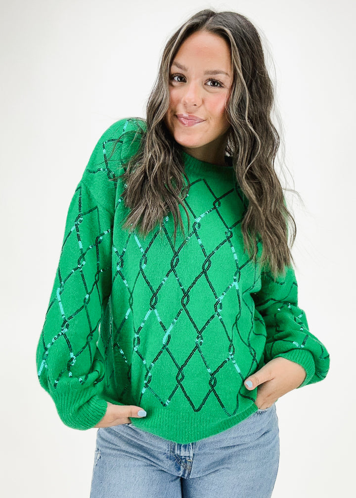 A woman wearing a green sweater with sequin-embellished diamond patterns and long sleeves, styled with light-wash jeans for a bold and cozy look.