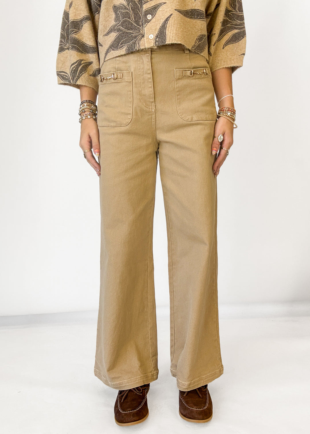 A person wearing tan wide-leg pants with front patch pockets, paired with a patterned beige and brown top and brown shoes, standing against a plain light background.