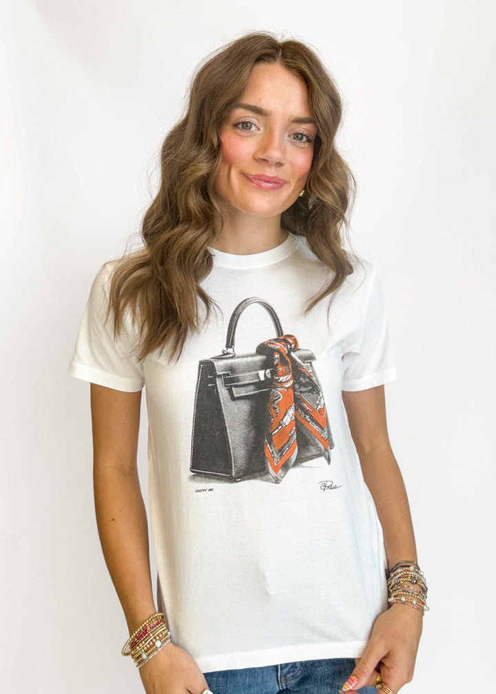A woman wearing a white graphic t-shirt featuring a black handbag with an orange and black patterned scarf, paired with blue jeans and gold bracelets, against a plain white background.