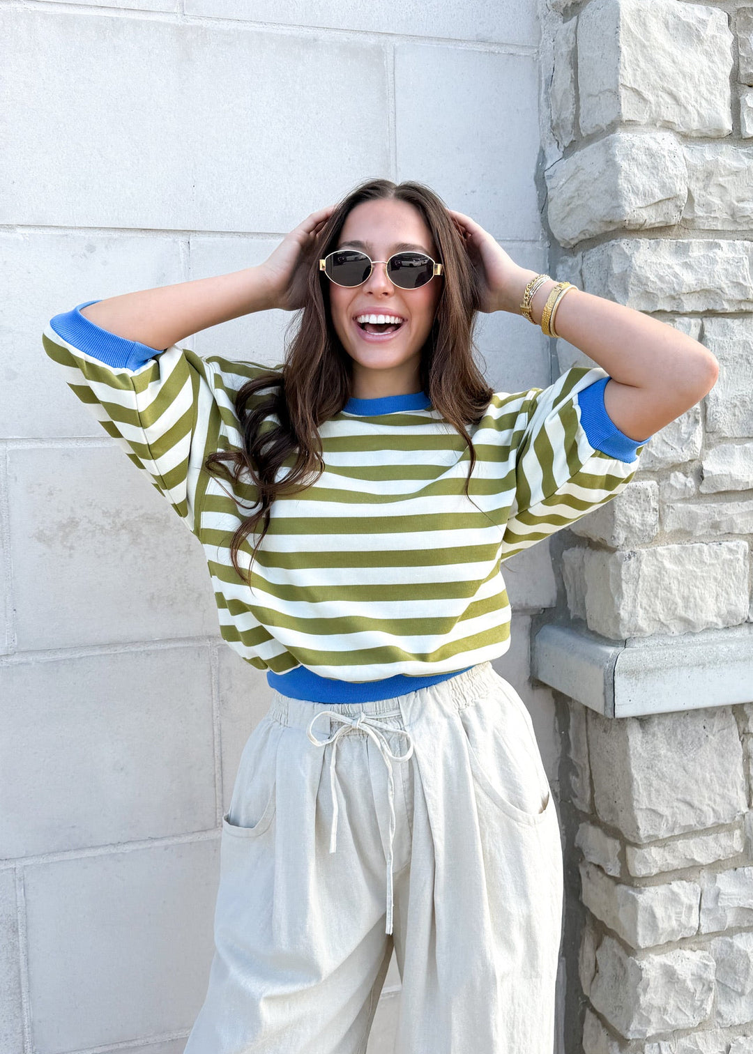 Woman wearing a green and white striped shirt and white pants leaning against a stone wall.