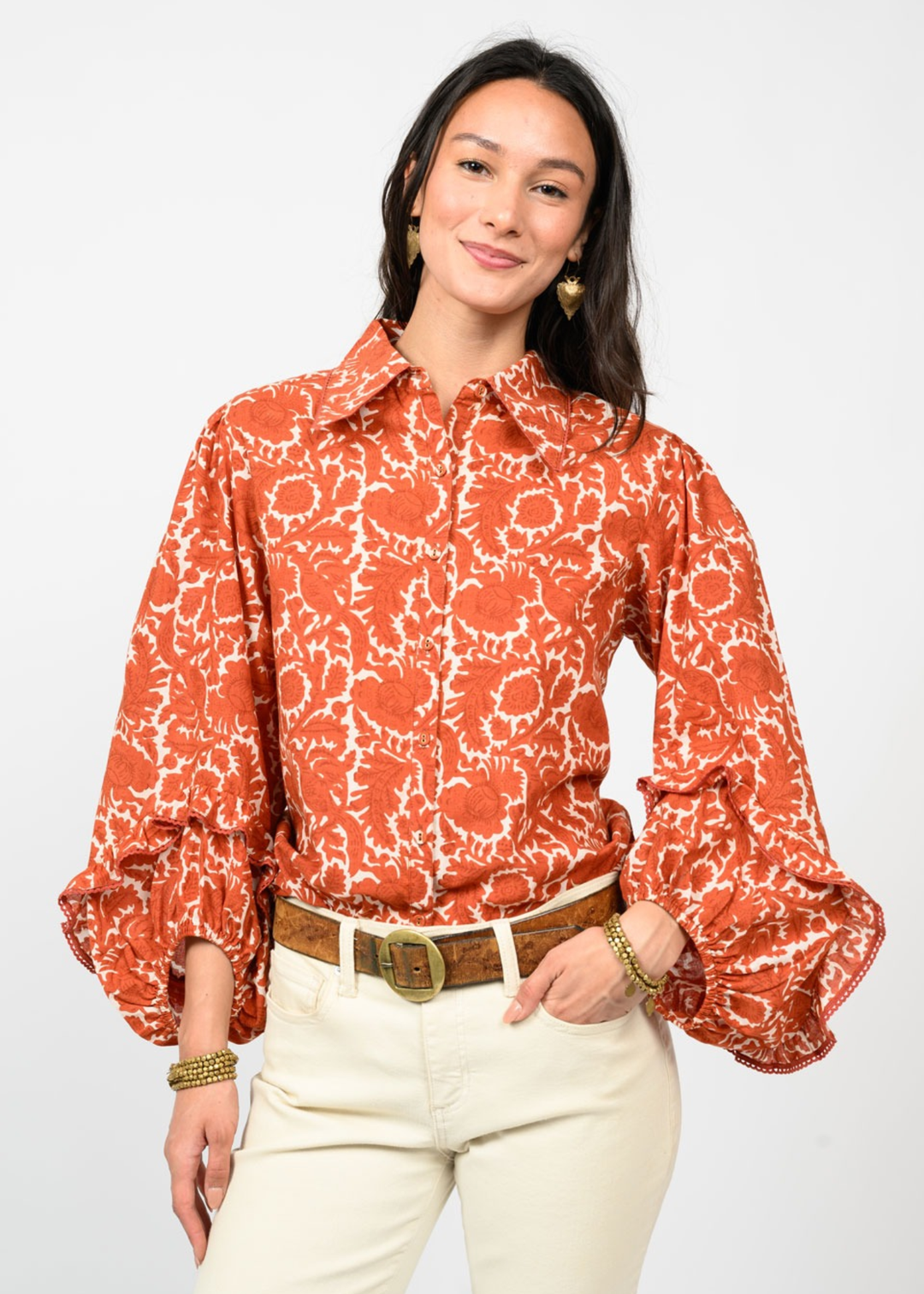 Woman wearing an orange floral patterned button down top with hand in pocket in front of a white background