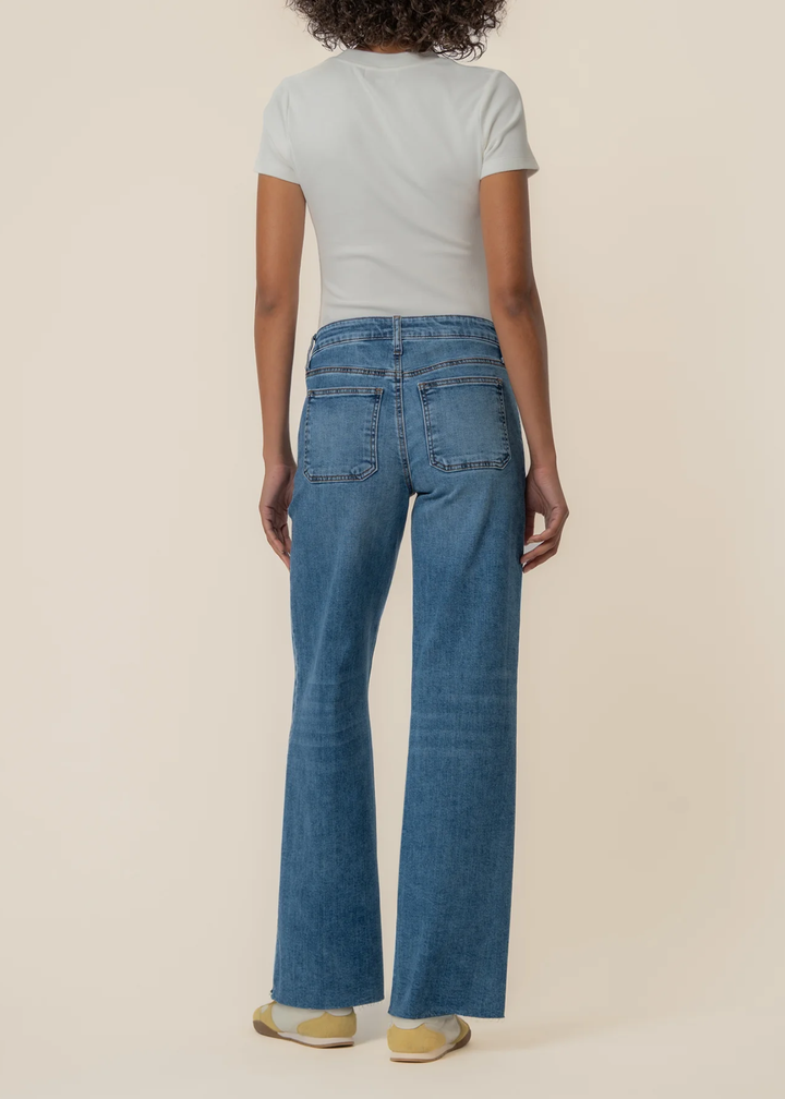 Woman turned around wearing blue wide leg jeans with a raw hem and front pockets against a tan background