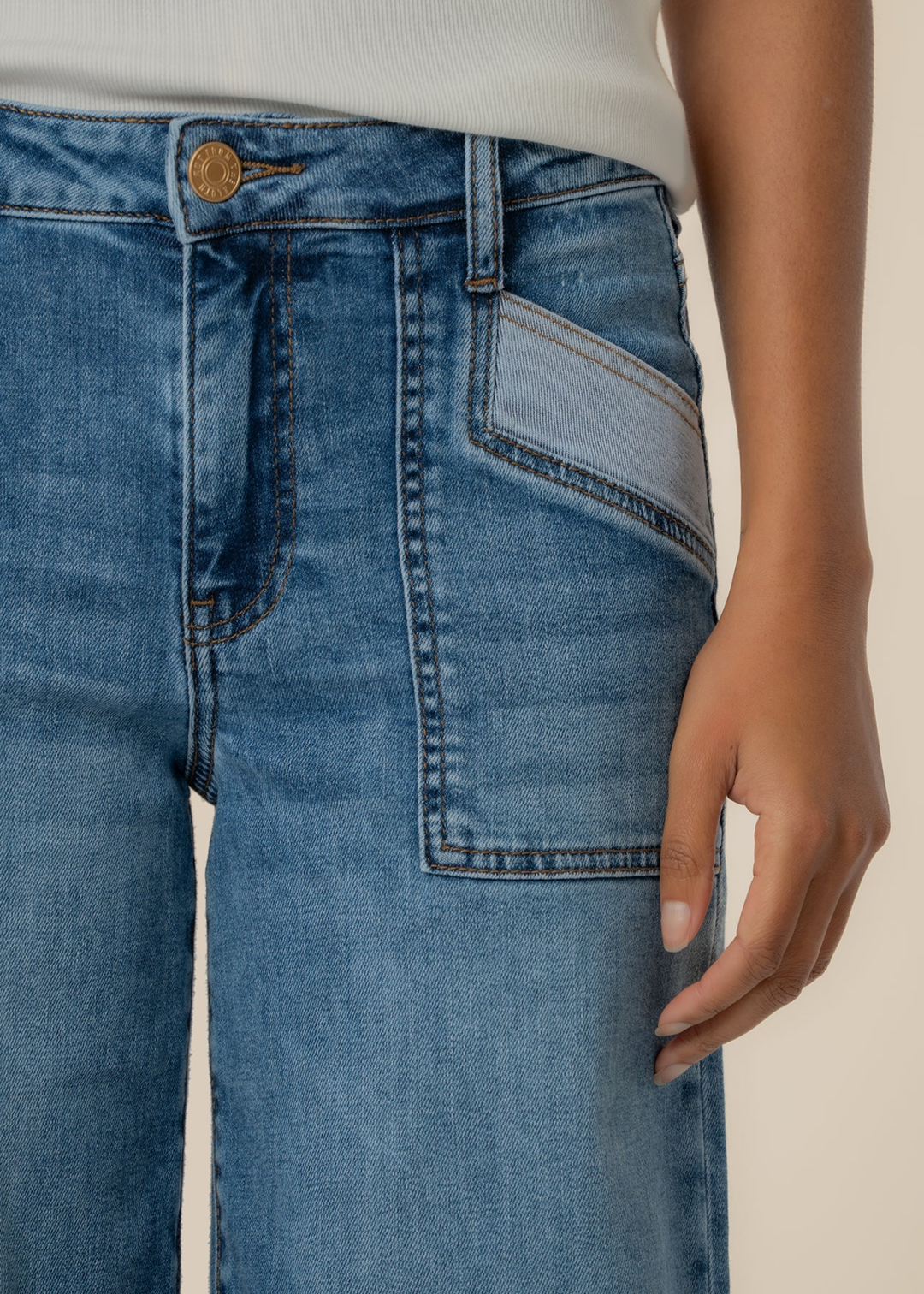 Close up of a woman wearing blue wide leg jeans with a raw hem and front pockets against a tan background