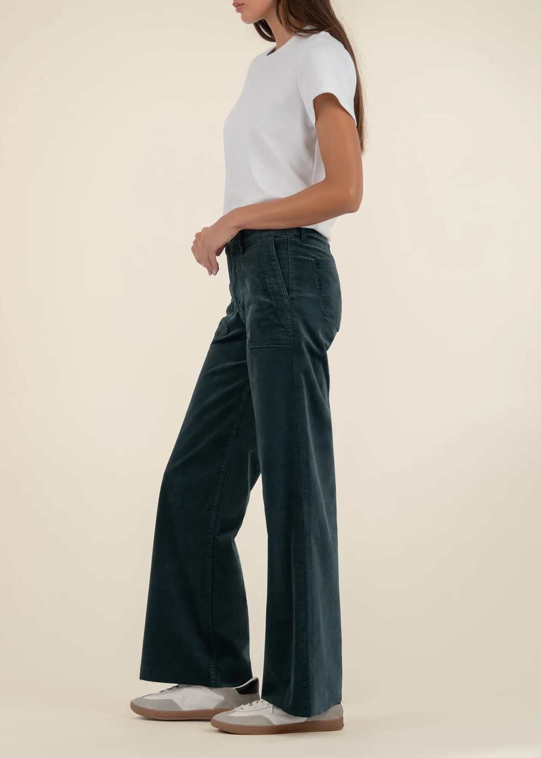 Woman turned to the side wearing teal velvet jeans with a white t-shirt and sneakers against a tan background