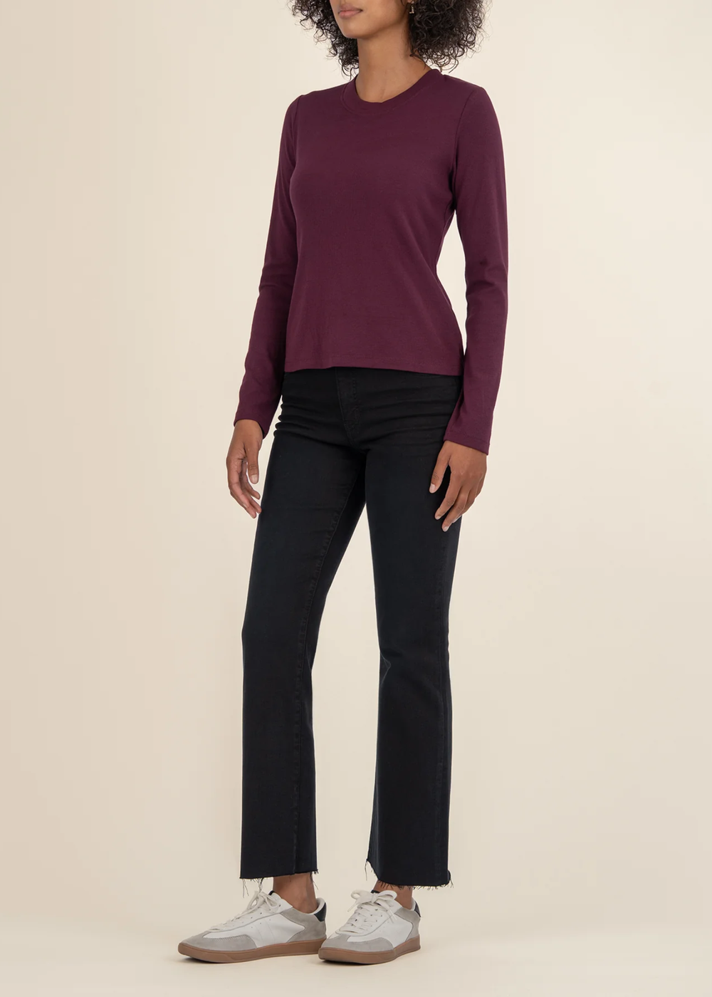 Woman turned to the side wearing a maroon long sleeve tee with black jeans and white sneakers in front of a white background