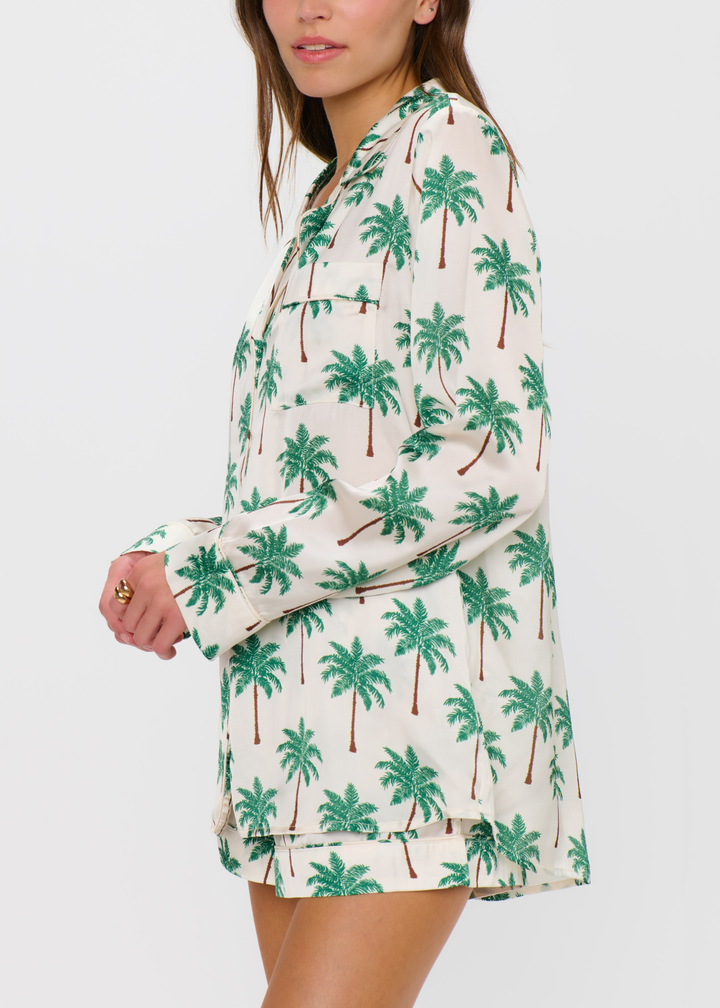 Woman turned to the side wearing a palm tree patterned pajama set with shorts and a long sleeve top against a white background