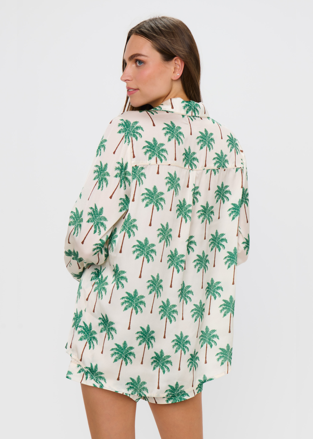 Woman turned around wearing a palm tree patterned pajama set with shorts and a long sleeve top against a white background