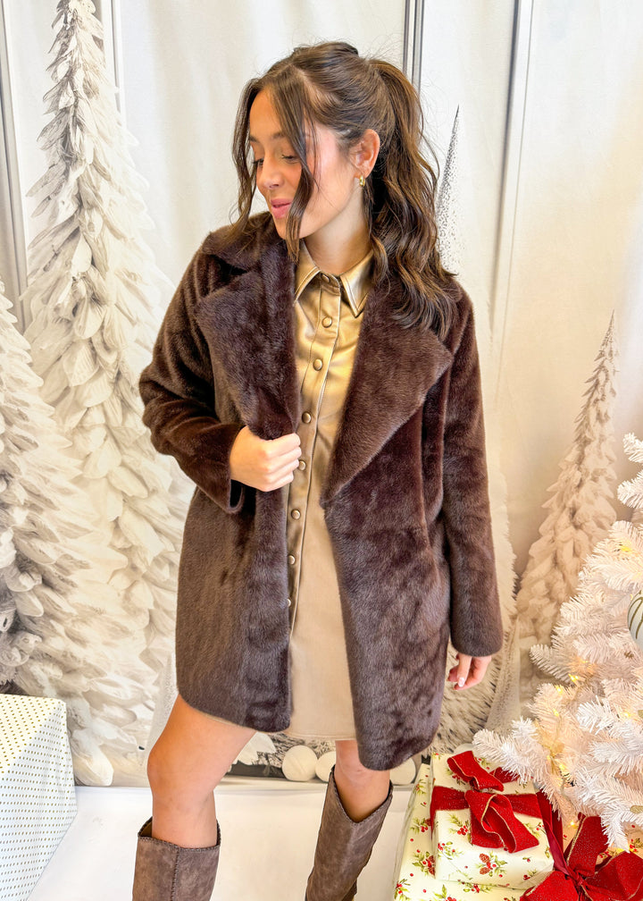 Woman wearing a brown faux fur coat in a festive setting with Christmas trees and presents.