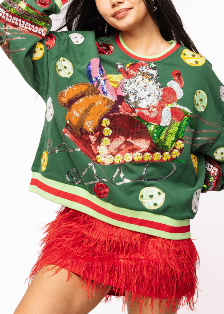 Woman wearing a green sweatshirt with sequin santa, sleigh, and ornament graphics with a red fringe skirt in front of a white background