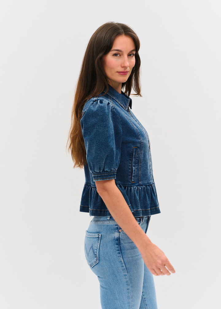 Woman turned to the side wearing a dark denim jacket with puff sleeves and a peplum design over a white shirt and light wash jeans