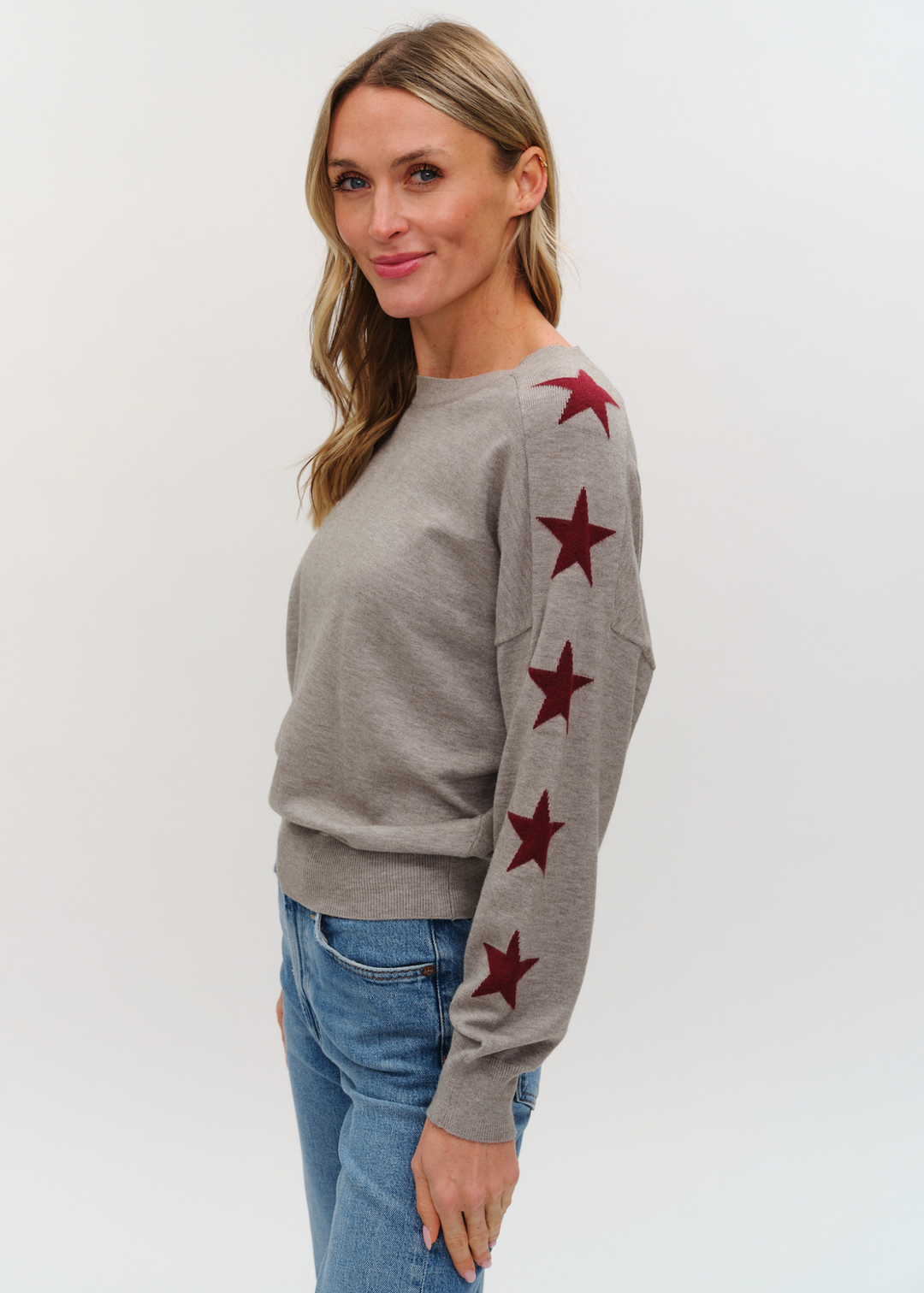 Woman wearing a grey sweater with maroon stars on the sleeves and jeans against a white background