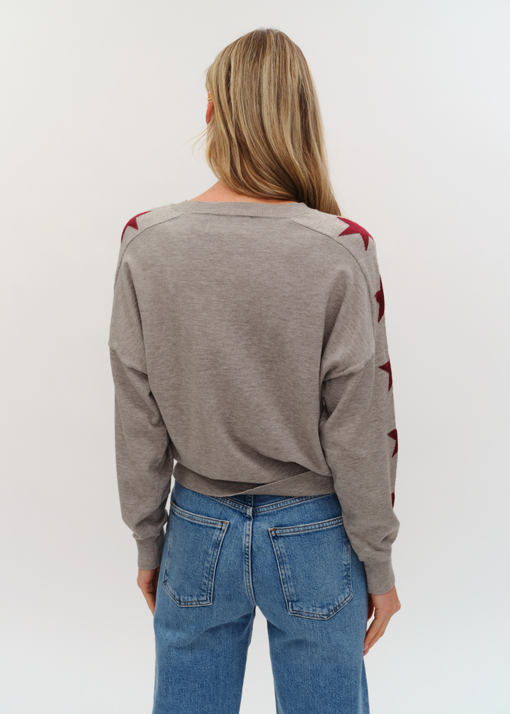 Woman turned around wearing a grey sweater with maroon stars on the sleeves and jeans against a white background