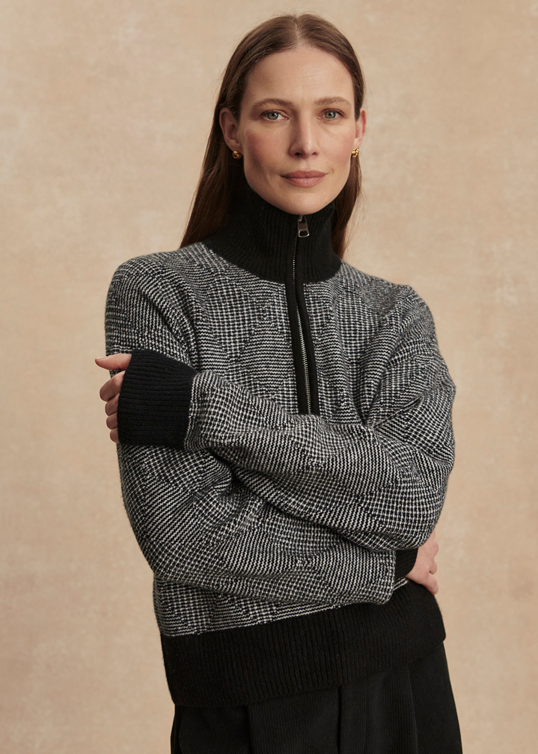 Woman with arms crossed wearing a black knit top with black pants against a tan background