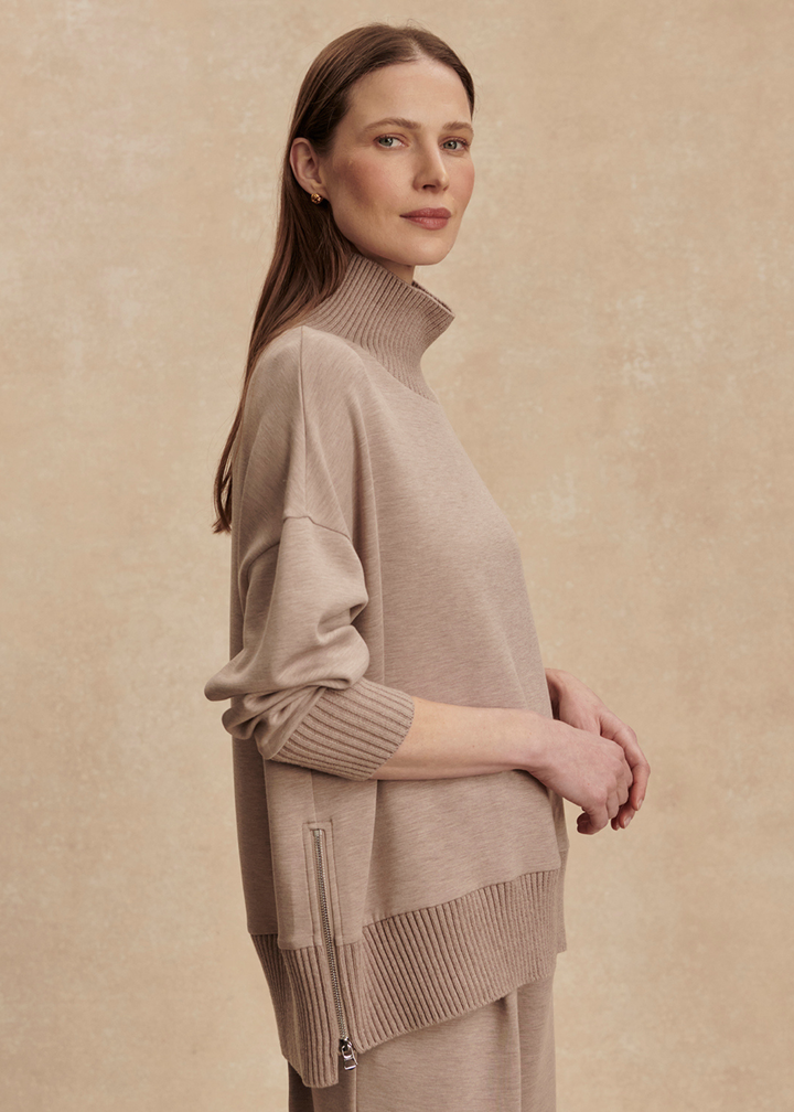 Woman turned to the side wearing a taupe high neck sweatshirt with taupe pants in front of a tan background