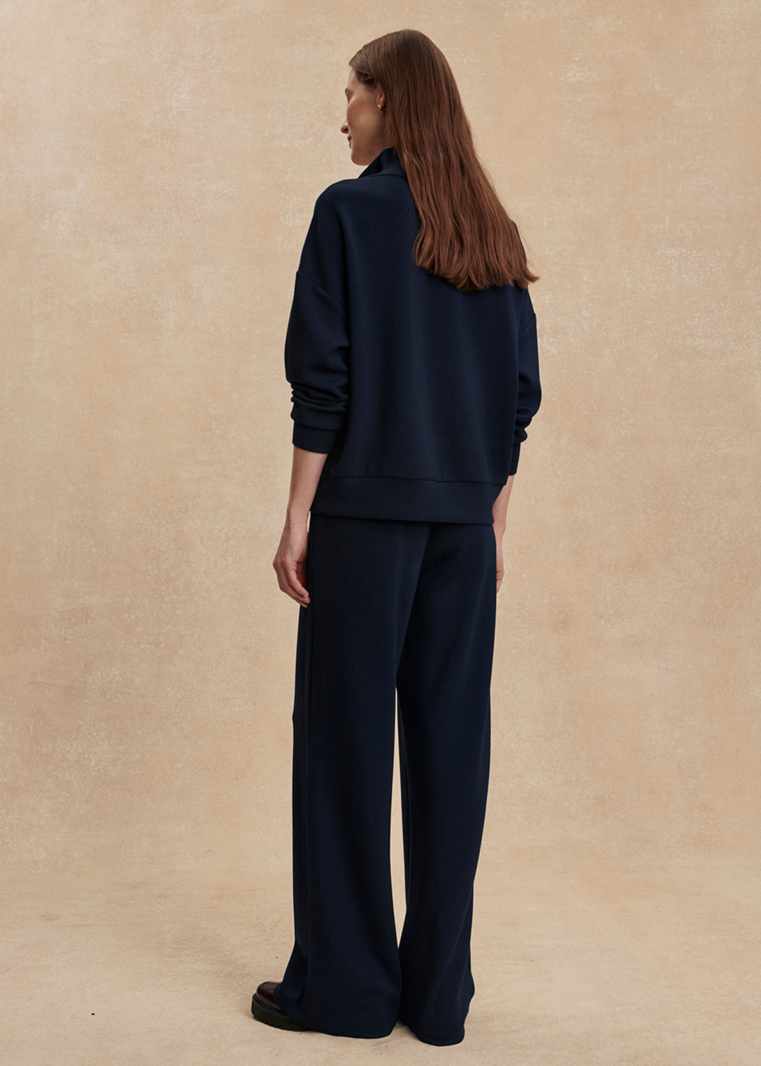 Woman turned around wearing wide leg navy sweatpants with a matching top in front of a tan background