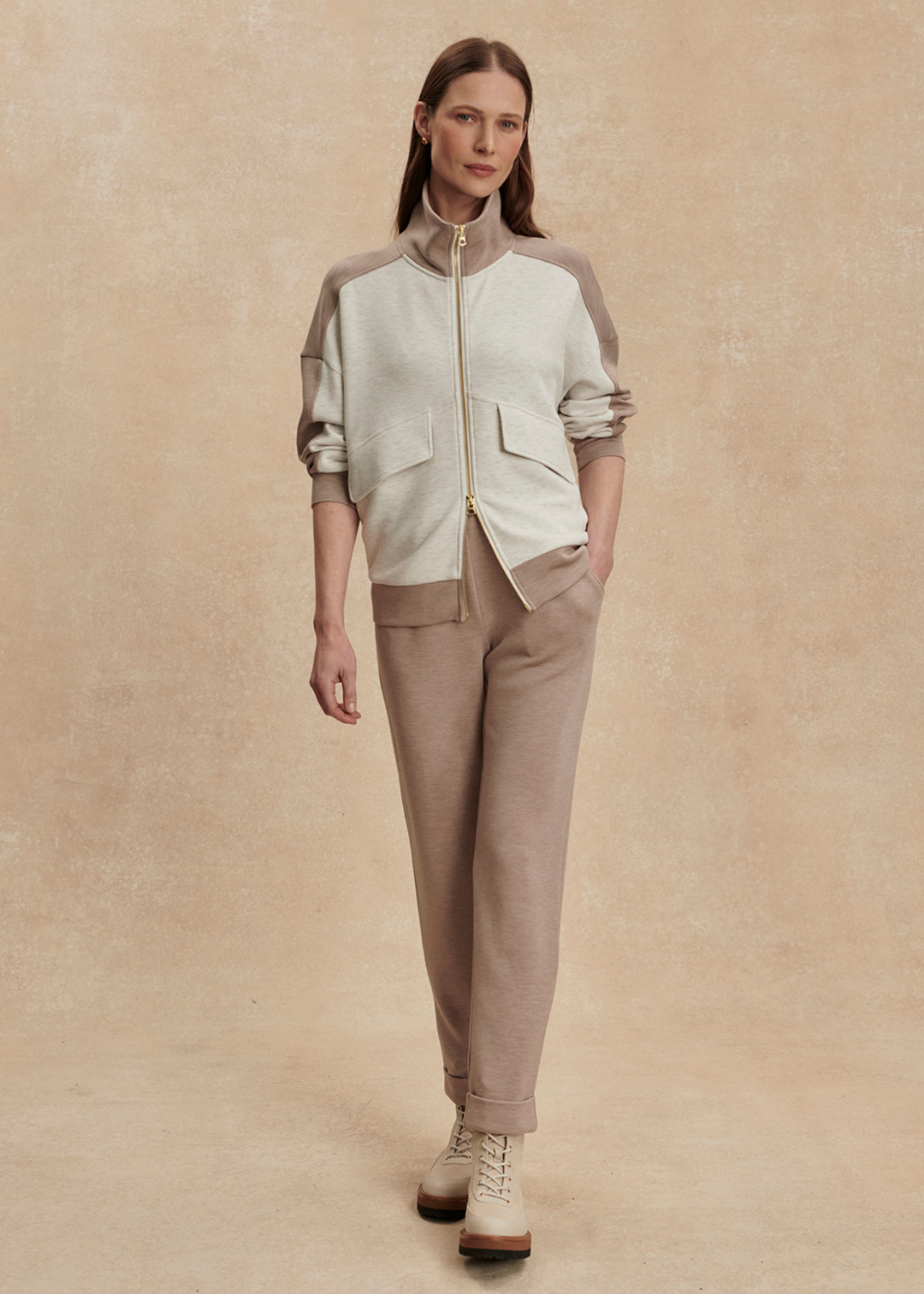 Woman with hand in pocket wearing a taupe and white zip up sweatshirt with taupe pants against a tan background 
