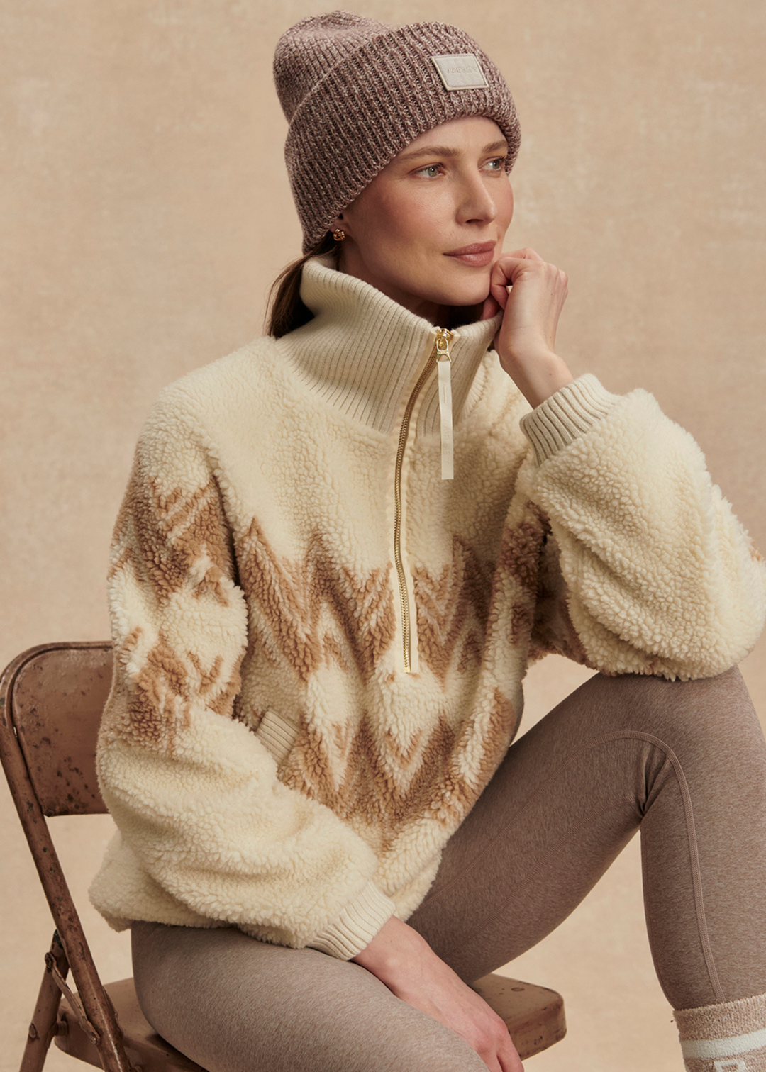 Woman sitting wearing a cream and brown half zip sherpa with a brown beanie and leggings