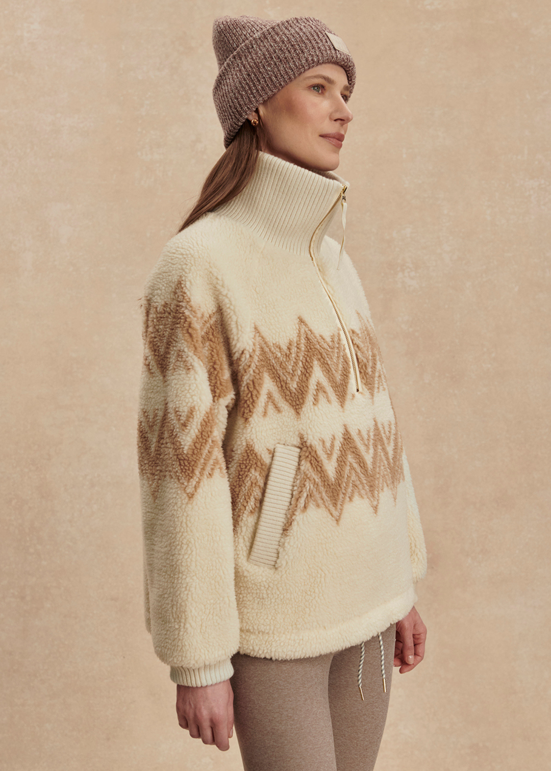 Woman turned to the side wearing a cream and brown half zip sherpa with a brown beanie and leggings