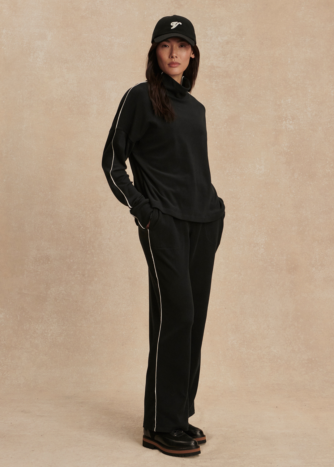Woman with hands in pockets wearing a black set with white trim against a tan background