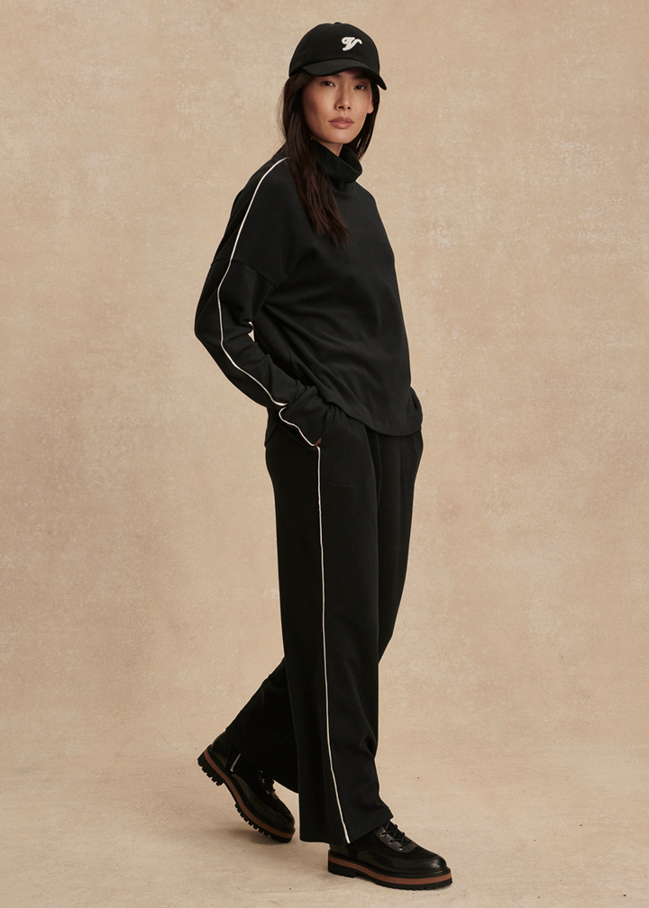 Woman with hand in pocket wearing a black set with white trim against a tan background