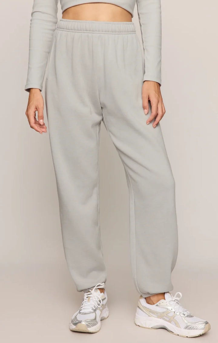 A person wearing light gray jogger sweatpants with an elastic waistband and cuffs, paired with white sneakers, shown from the waist down against a plain light beige studio background.