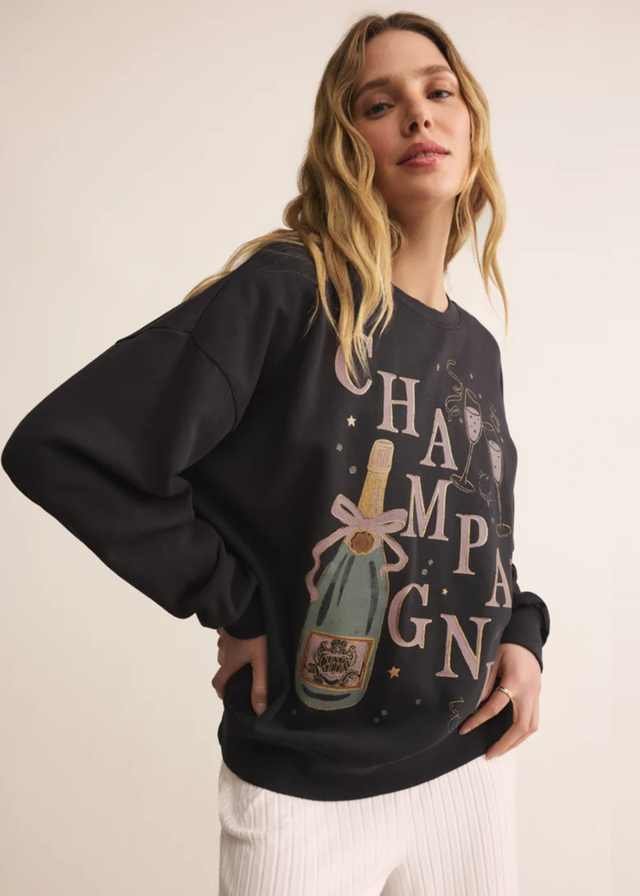 Woman with hand on hip wearing a grey sweatshirt with a champagne graphic and white pants